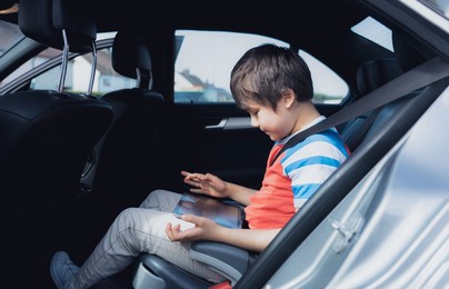 cinematic portrait happy young boy using a tablet computer while sitting in the back passenger seat with a safety belt, child typing on smart pad,school kid traveling to school by car.back to school
