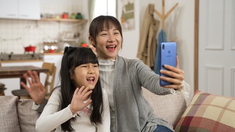 cheerful asian mom and daughter waving hands while chatting online with friends through video conferencing app in the living room at home