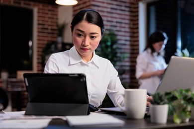 asian businesswoman with modern tablet reviewing accounting documentation in office workspace. executive manager working on electronic touchscreen device while developing marketing strategy.