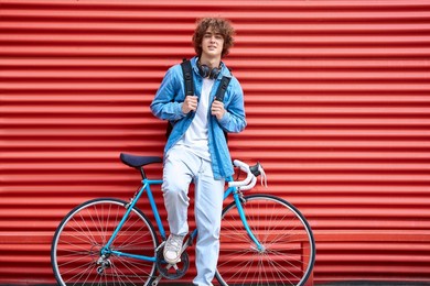 stylish young male in casual clothes keeping hands on backpack straps while leaning on red metal wall and bike and looking at camera