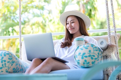 portrait image of a beautiful young asian woman using laptop computer for remote working or studying online while drinking coffee and sitting on swing sofa