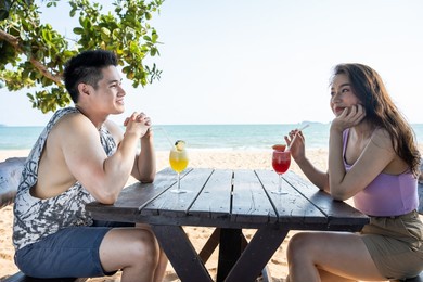 asian young man and woman couple drinking juice on the beach together. attractive new marriage people sitting on dinner table at seaside enjoy holiday vacation honeymoon trip in tropical sea island.
