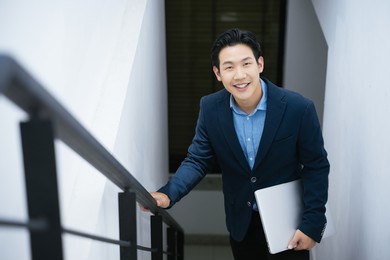 portrait asian businessman smiling confidently in office stairs with holding laptop.