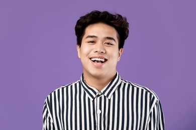 portrait of excited young asian male laughing wearing casual striped shirt posing standing isolated over purple violet studio background. happy adult person smiling looking at camera with open mouth