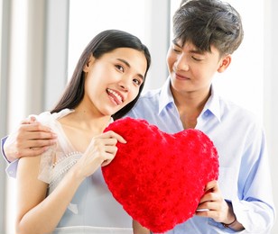 asian man and woman lovers cuddle holding together with heart shape red pillow.