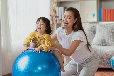 happy asian child having fun with gymnastic ball, doing exercises with mother at home