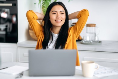 happy chinese girl, freelancer, working from home, sits in the kitchen at the workplace, takes a break from work, puts her hands behind her head, looks to the side, dreams of rest, thinks, smiles