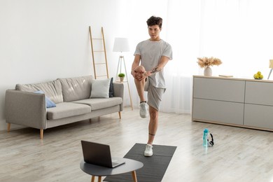 home workout routine. focused fit asian guy lifting leg up to chest stretching hamstring muscles, doing standing knee-to-chest exercise, using pc in living room indoors. healthy lifestyle concept