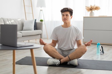 online yoga lesson. calm focused asian guy meditating alone looking at pc screen, watching online tutorial sitting on floor yoga mat in living room, using laptop on tea coffee table, selective focus