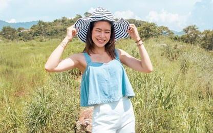 beautiful cheerful asian female traveler wearing casual fashionable clothes, hat, standing in outdoor green field or park, smiling with happiness while traveling in summer holiday vacation