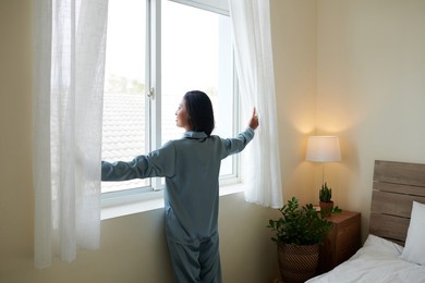 smiling young woman in pajama opening curtains and looking outside