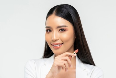 friendly young asian female with lipstick smiling and looking at camera during makeup application against white background