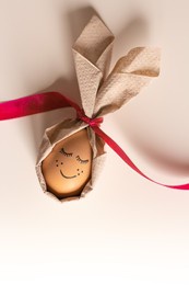beautiful easter egg with a painted smile, eyelashes and freckles in bunny ears from a napkin on a white background. easter ukraine 2022. cool egg for easter with a painted face and a red ribbon