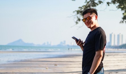a young man with sunglasses playing mobile phone by the sea on vacation with free space