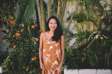 front view of smiling asian girl posing in yard with tropical plants on bali island. cropped image of young beautiful brunette indonesian woman in dress looking at camera at sunny day