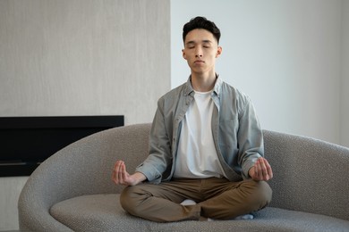 focused asian man meditates in lotus pose breathing deep and slowly. young male person in casual clothes sits on comfortable coach relaxing at home