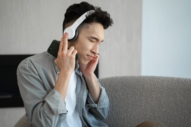 asian man in headphones listen to dynamic music shaking head sitting on sofa. young guy in casual clothes enjoys weekend holding phone at home