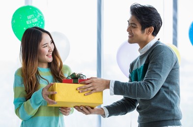 two asian lovers enjoying gift exchange together with happy faces at romantic indoor party decorated by fancy balloon to celebrate ecstatic love in winter