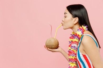 side view young woman of asian ethnicity in striped one-piece swimsuit hawaii lei drink coconut straw cocktail juice isolated on plain pastel pink background. summer vacation sea rest sun tan concept