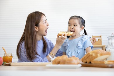 mother feeding bread toast to cute playful little girl or her daughter in the kitchen