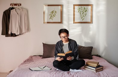 serious young asian student in eyeglasses sitting with crossed legs on bed and reading literature before exam