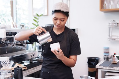 smiling asian barista young woman pouring hot black coffee into paper cup for according to the customer's order at counter bar in coffee shop.