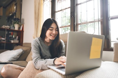 young adult southeast asian woman stay and working at home using laptop computer on sofa. people lifestyle with internet online technology. window light on day background. 