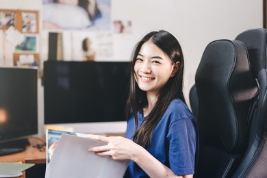 portrait of smile young adult southeast asian woman stay at home office reading a book and relax. her longhair and wearing blue shirt. beauty face looking at camera.