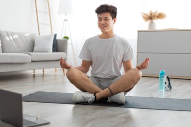 keep calm. happy sporty asian man meditating with trainer online via laptop connection using pc in living room. healthy guy sitting on the floor yoga mat in lotus position, looking at computer screen