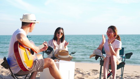 happy young people enjoying with music at the beach, young man playing guitar and singing as his friends clapping and laughing, having fun on the beach picnic in summer holidays, vacation.