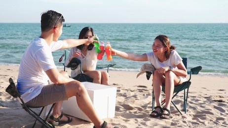 happy young people clinking bottles and drinking on the beach, friends having fun with beach picnic party in summer camping, vacation and lifestyle concept.
