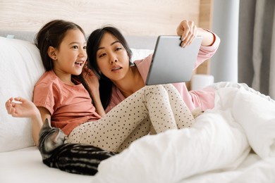 asian mother and daughter using tablet computer while sitting on bed at home