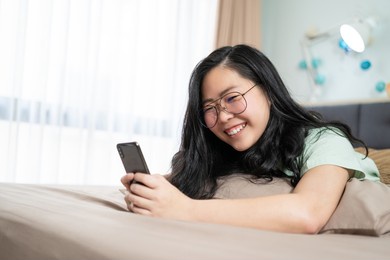 beautiful glasses asian woman lies on her stomach and plays mobile on the bed in her bedroom with a pastel green - brown color theme.