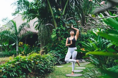 asian girl practicing yoga in tree pose outdoor. concept of harmony and mental health. young beautiful athletic woman wearing sportswear and barefoot in tropical garden. bali island. daytime