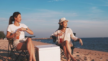 two teenage girls sitting on beach clinking bottles and drinking soda, group of friends enjoying drinks and playing guitar in camping beach at sunset, vacation and lifestyle concept.