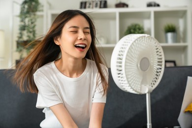 young smiling asian woman sitting on couch and turned on fan to cool herself with suffers from too hot weather in living room.