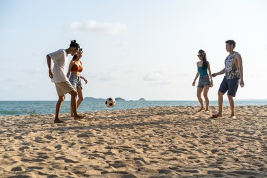 group of asian young man and woman play soccer on the beach together. attractive friend traveler feel happy and relax, having a football game while travel for holiday vacation in tropical sea island.