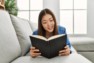 young chinese girl reading book lying on the sofa at home.