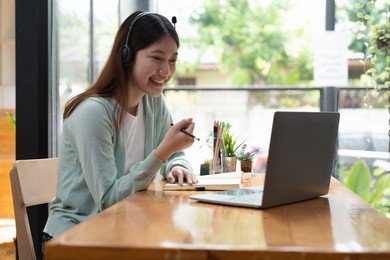 smiling girl student wear wireless headphone study online with teacher, happy young asian woman learn language listen lecture watch webinar write notes look at laptop sit in cafe, distant education