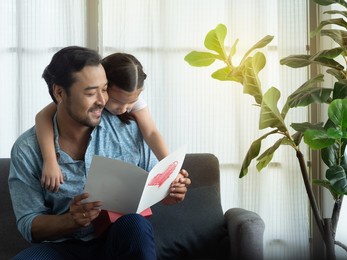 handsome asian father reading card while cute little daughter hugging dad's from the back. father's day or birthday gift and  card.