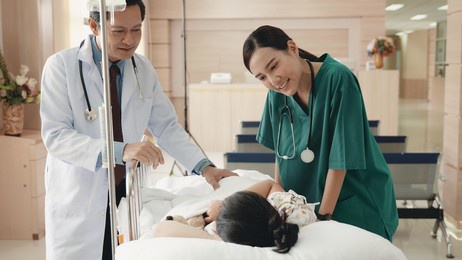 cute little sick girl patient lying in hospital bed talking with smiling doctor and nurse standing at hospital, medicine healthcare and pandemic, friendly doctor concept