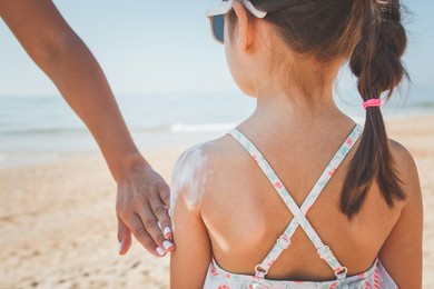 mom smears her daughter with sunscreen on the beach in summer, the girl is standing with her back, her face is not visible, the concept of safe tanning.