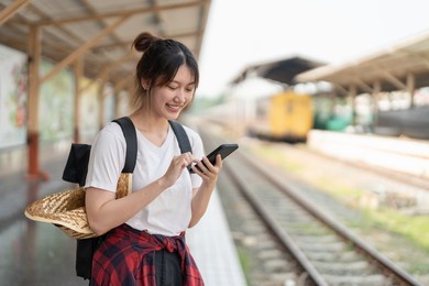 beautiful young asian woman with a backpack using mobile phone while standing near the railroad train on the platform. cheap travel summer concept