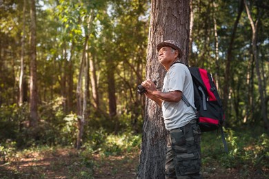 an elderly asian man and backpack watching birds with binoculars on trees in the forest. old man hiking on vacation.