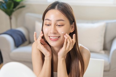 fresh healthy skin, beautiful smile of asian young woman, girl looking at mirror, applying moisturizer on her face, putting cream treatment before makeup cosmetic routine at home. facial beauty.
