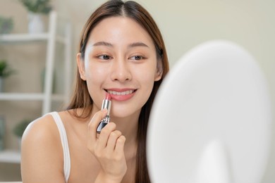 skin care cosmetics concept, beautiful asian young woman, girl make up face by applying lipstick, lips balm on her mouth, looking at the mirror at home. female look with natural fashion style.