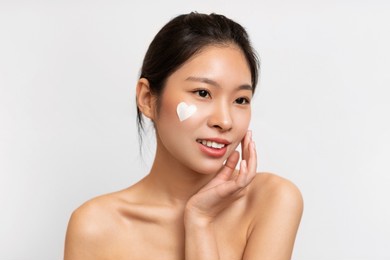 facial skincare concept. portrait of attractive dreamy young asian lady with heart-shaped face cream on cheek posing on white studio background wall, touching face. skin care cosmetics advertisement