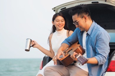 happy asian couple lover having stop off by the sea and sitting in hatchback  ,they are  drinking coffee playing guitar together.