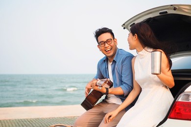 happy asian couple lover having stop off by the sea and sitting in hatchback  ,they are  drinking coffee playing guitar together.