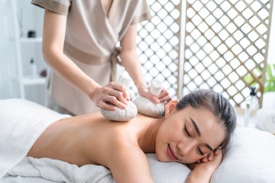 asian beautiful woman enjoy thai hot compress massage with herbal bags. attractive girl lying on massage table, feel happy and relax during back physiotherapy from masseuse for body care in spa salon.